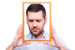 Depressive portrait. Portrait of frustrated young man in shirt holding picture frame in front of his face while standing against white background
